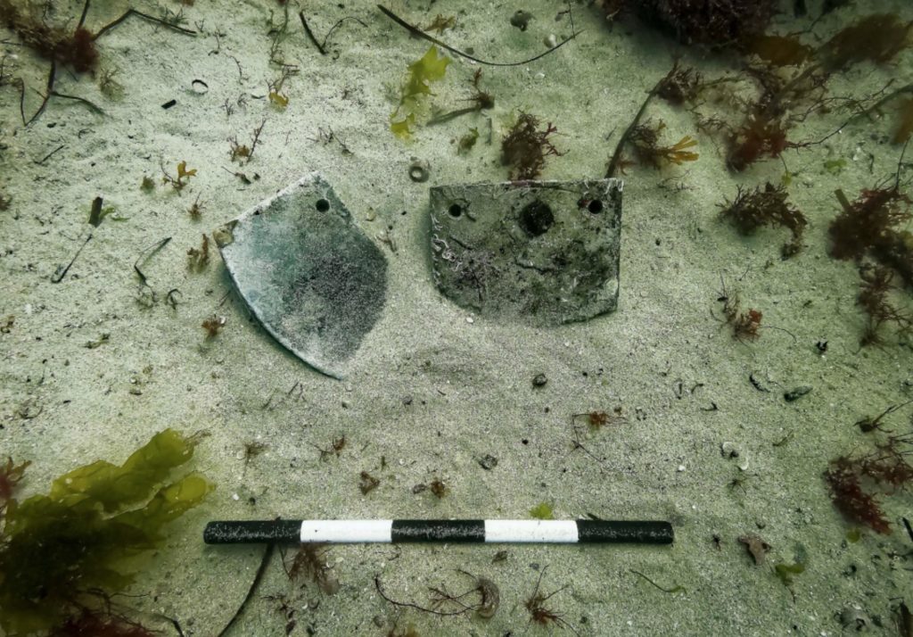 Underwater photo of two grey lead objects laying on the seabed in the sand. At the bottom of the photograph is a black and white scale measuring half a metre.