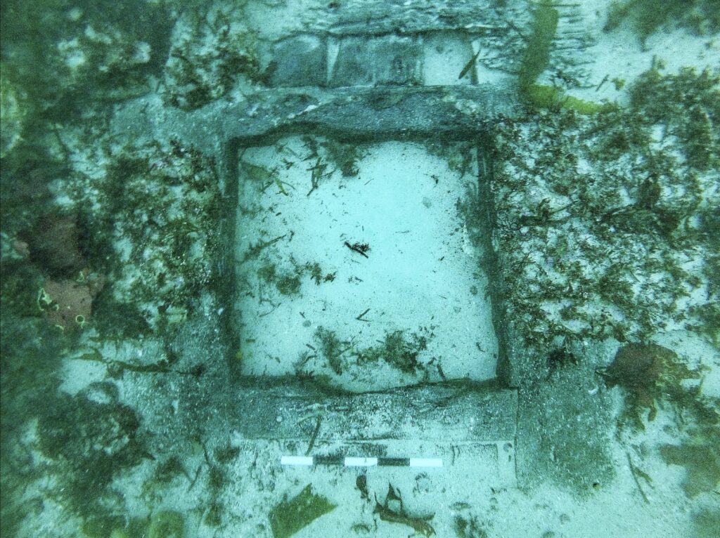 The gunport, located on the seabed, is a square opening framed by the remnants of the ship's structure, which is encrusted with marine growth. The edges of the gunport are outlined by preserved timber elements, partially buried in sediment. On the top left timber of the gunport can be observed a curved depression, created by the gun repeatedly striking it when fired. Seaweed and sand partially cover the surrounding area, blending with the wreck site.

A black-and-white scale bar is positioned at the bottom centre of the frame.